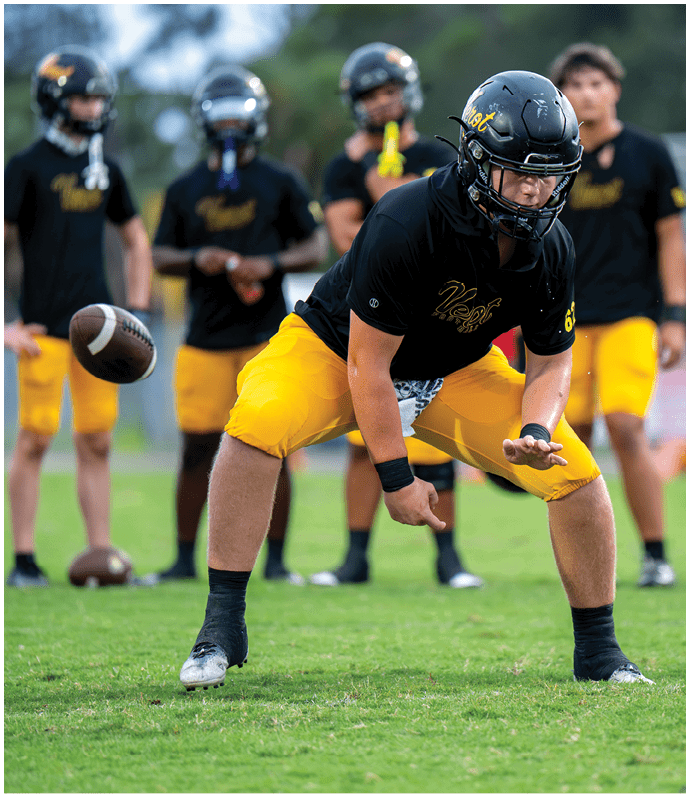 A person wearing a black helmet, black shirt, and yellow shorts is preparing to catch a football while several others in similar attire stand in the background on a grassy field. Descripci n generada con IA