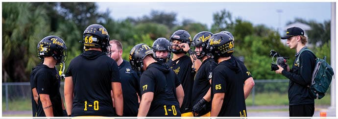 A group of people wearing black football helmets and uniforms are gathered on a field while another person with a backpack films them with a camera. Descripci n generada con IA