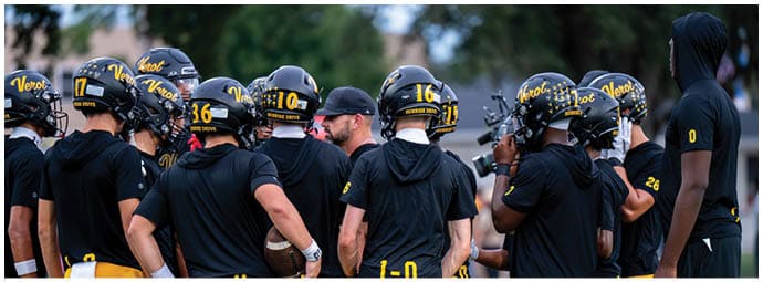 A group of people wearing black and yellow football helmets and athletic clothing are gathered closely outdoors, seemingly listening to a coach or leader. Descripci n generada con IA