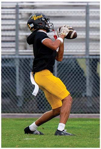 A person wearing a black helmet, black shirt, and yellow pants is catching a football on a grassy field with empty bleachers and a chain-link fence in the background. Descripci n generada con IA