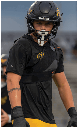 A person wearing a black football helmet and black and yellow athletic gear stands in the foreground while another person in similar attire holds a football in the background on a sports field. Descripci n generada con IA