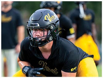 A person wearing a black football helmet and black sports jersey is focused and crouched in an athletic stance on a field. Descripci n generada con IA