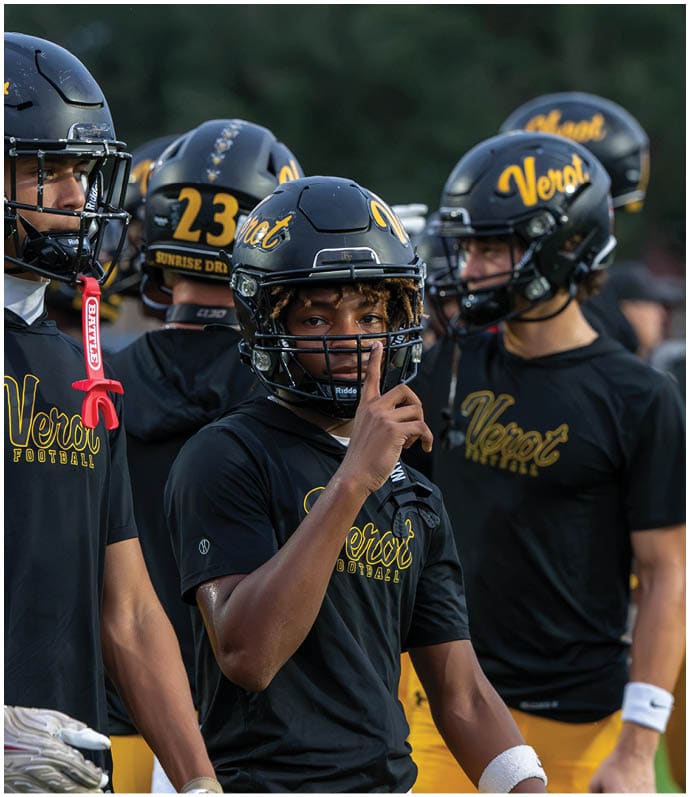 A person wearing a black football helmet and jersey with “Verot" written on them holds a finger to their lips while standing among other similarly dressed people. Descripci n generada con IA
