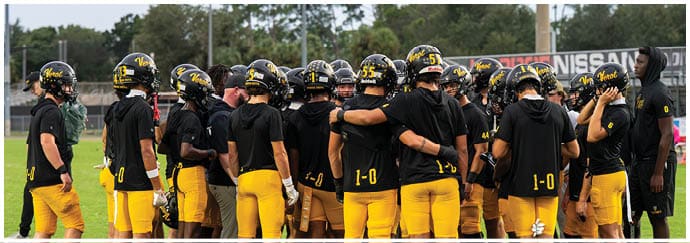 A group of people wearing black shirts, yellow pants, and helmets are gathered closely together on a sports field, appearing to be in a team huddle. Descripci n generada con IA