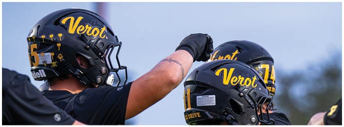 Three people wearing black football helmets with yellow “Verot" lettering are engaged in a team huddle or celebration outdoors. Descripci n generada con IA