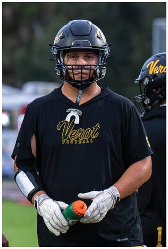 A person wearing a black football helmet and a black sleeveless shirt with the word “Verot Football" written in yellow stands outdoors. Descripci n generada con IA