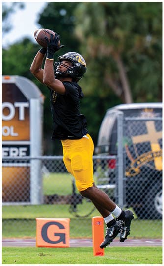 A person wearing a black helmet, black shirt, and yellow pants is jumping to catch a football near an orange marker on a sports field. Descripci n generada con IA