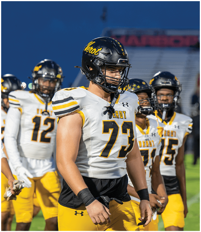 A group of people wearing football helmets and white jerseys with yellow and black accents stand together on a field during dusk. Descripci n generada con IA