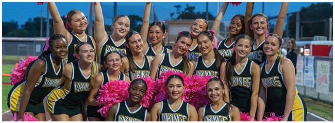 A group of people in cheerleading uniforms holding pink pom-poms raised in the air, smiling and posing together outdoors at dusk. Descripci n generada con IA