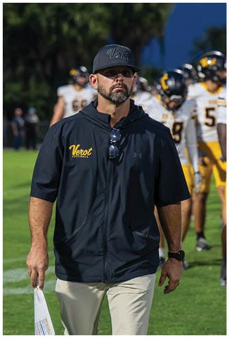 A person wearing a dark jacket and cap with “Verot Football" written on them is walking on a football field holding a paper, with several people in football uniforms standing in the background. Descripci n generada con IA