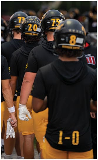 A group of people wearing black shirts, yellow pants, and black helmets with numbers and the words “Sunrise Drive" stand closely together, some holding white gloves. Descripci n generada con IA