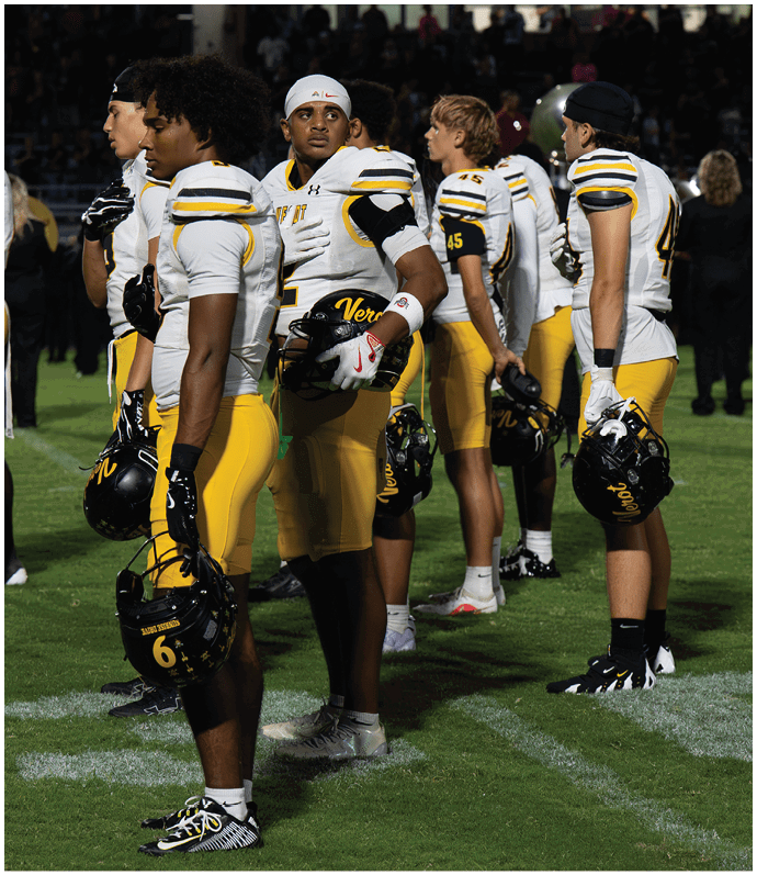 A group of adults in white and yellow football uniforms stand on a grassy field holding helmets, appearing attentive and ready for a game. Descripci n generada con IA