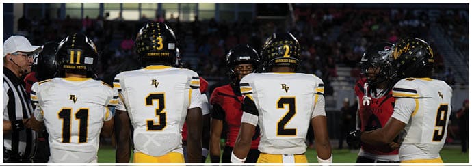 Four people in football uniforms with numbers on their backs stand on a field facing away from the camera while two others in different uniforms stand facing them. Descripci n generada con IA