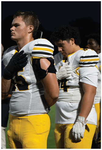 An adult wearing a white and yellow football uniform stands with a hand over their chest during what appears to be a pre-game or national anthem ceremony on a field at night. Descripci n generada con IA