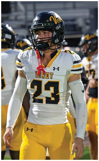 A person wearing a football uniform with the number 23 and a helmet stands on a field with other players in the background. Descripci n generada con IA