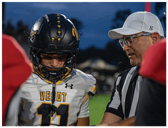 A person wearing a football helmet and uniform stands next to a person in a referee uniform with a headset, both appearing focused during a game at dusk. Descripci n generada con IA