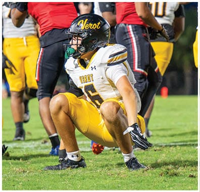 Two people in yellow and white football uniforms with helmets are crouched on a grassy field while others stand in the background. Descripci n generada con IA