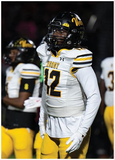 A person wearing a black helmet and a white football uniform with yellow and black stripes and the number 12 stands on a field. Descripci n generada con IA