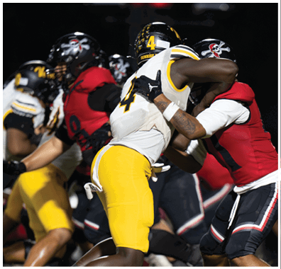 Two people in football uniforms are engaged in a physical play during a game, with one wearing a white and yellow uniform and the other in a red and black uniform. Descripci n generada con IA