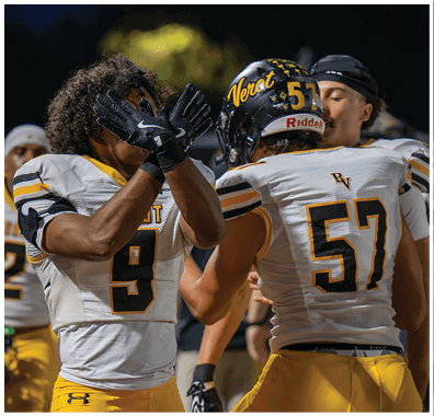 Two football players in white and yellow uniforms are interacting on the field, with one player crossing their gloved hands in front of their face. Descripci n generada con IA