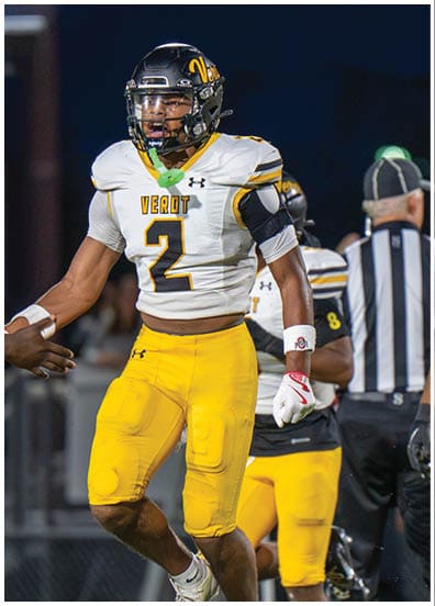 A person wearing a football uniform with the number 2 and the word “VEROT" on the jersey is jumping and giving a handshake to another person on a football field during a game. Descripci n generada con IA