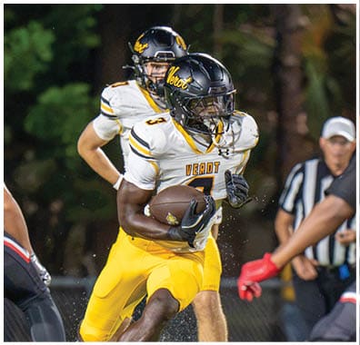 A person in yellow football gear is running with the ball while being pursued by players in red and black uniforms on a grassy field during a nighttime game. Descripci n generada con IA