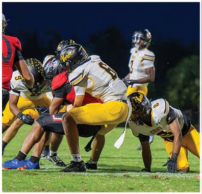 People in football uniforms are positioned on a grassy field at night, preparing for a play with some crouched and others standing. Descripci n generada con IA