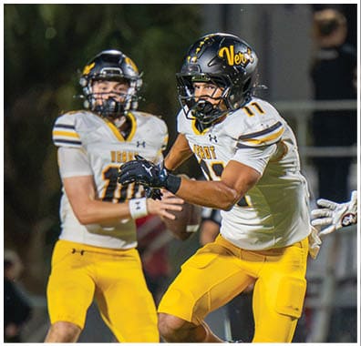 Two people in yellow and white football uniforms are actively playing on a field while a person in a red and black uniform approaches them. Descripci n generada con IA
