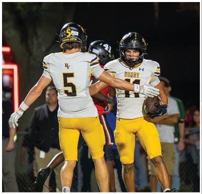 Two football players in yellow pants and white jerseys celebrate on the field during a nighttime game with spectators in the background. Descripci n generada con IA