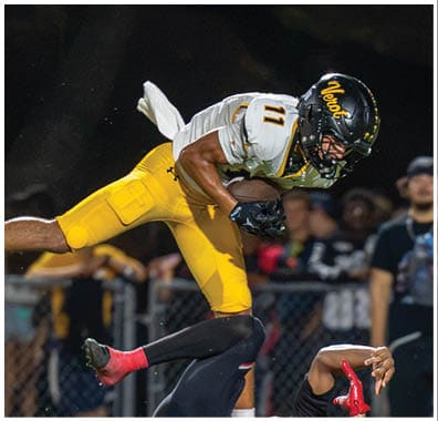 A person in yellow sports gear leaps over another person in black and red sports gear who is lying on the grass during a nighttime football game. Descripci n generada con IA