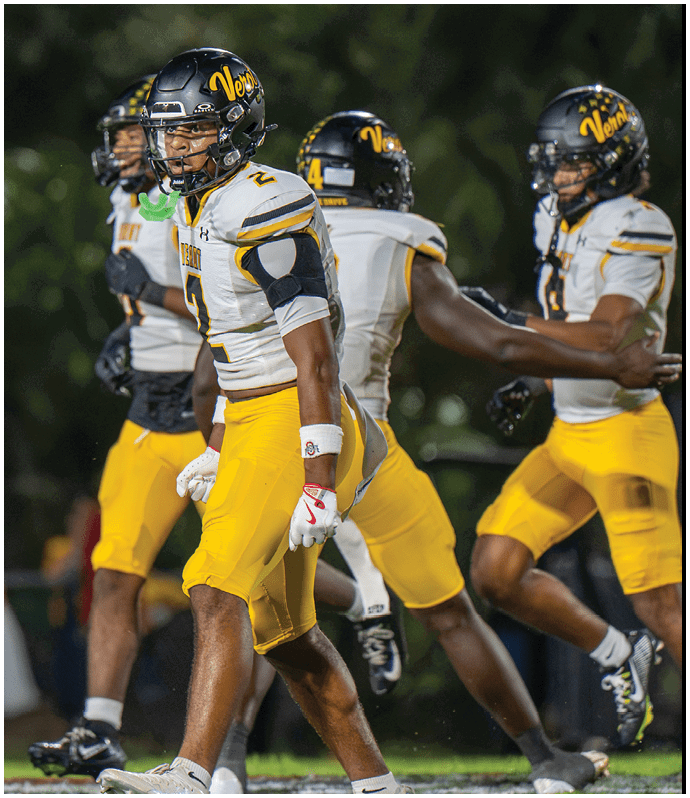 Three people wearing yellow football pants and white jerseys with black helmets are on a football field, with one person in the foreground appearing focused and ready for action. Descripci n generada con IA