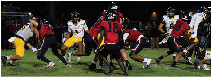 People wearing red and white football uniforms are actively engaged in a play on a grassy field under stadium lights with spectators in the background. Descripci n generada con IA