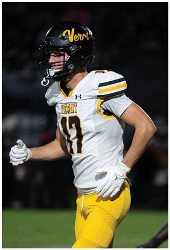 A person wearing a white football jersey with yellow and black stripes and a black helmet stands with one arm bent at the elbow. Descripci n generada con IA