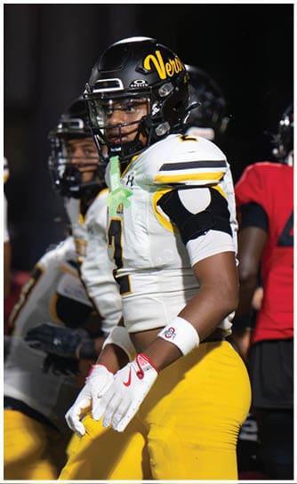 A person wearing a black football helmet and white and yellow football uniform stands on a field with other players in the background. Descripci n generada con IA