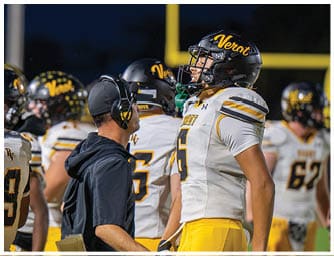 A person wearing a headset and cap is facing and talking to a football player in a helmet and uniform with the word “Vent" on it during a nighttime game. Descripci n generada con IA