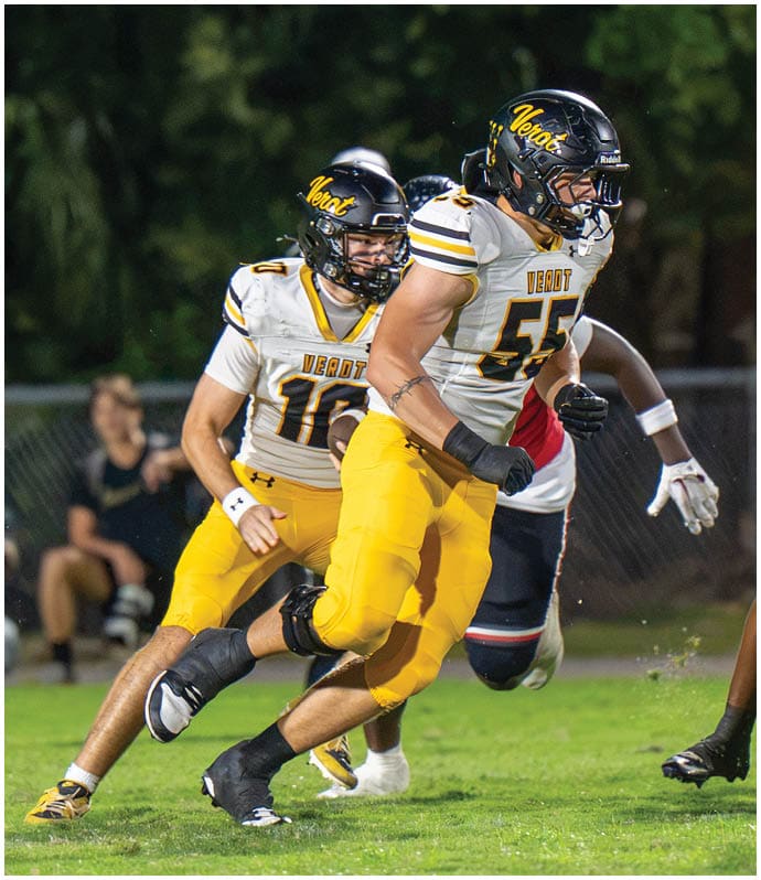 Two people in yellow and white football uniforms with black helmets are running on a grassy field during a game. Descripci n generada con IA