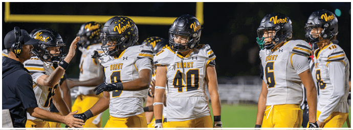 A group of football players in yellow and white uniforms with helmets are standing on a field at night, with one player shaking hands with a coach. Descripci n generada con IA