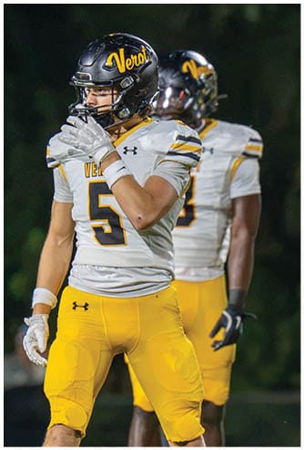 Two people wearing yellow football pants and white jerseys with black helmets stand on a field, with one person in the foreground holding a hand near their face. Descripci n generada con IA