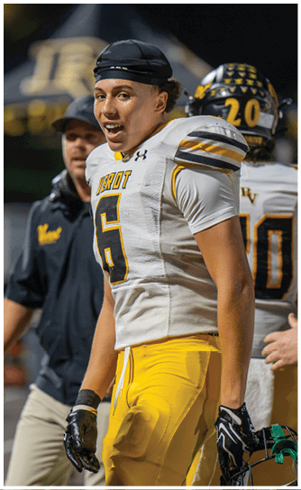 A person wearing a football uniform with the number six is standing on a field, smiling, with another person in sportswear and a helmeted player in the background. Descripci n generada con IA
