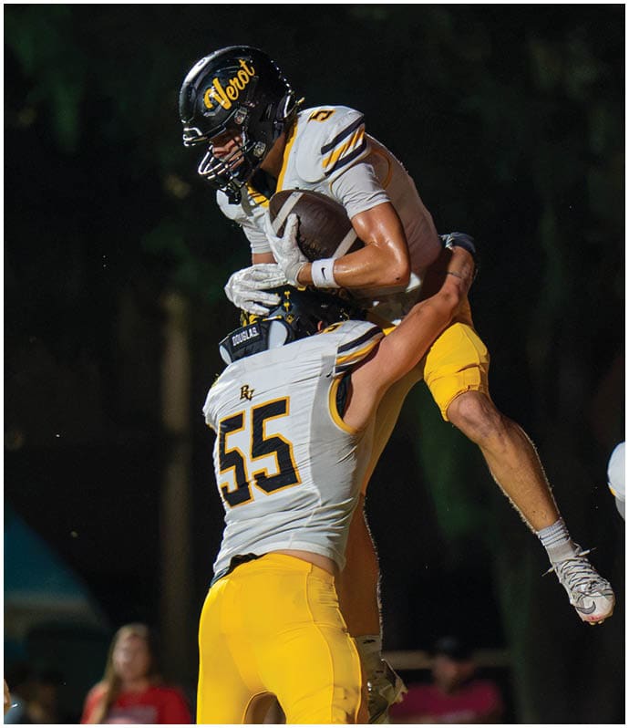 Two people in football uniforms celebrate on the field with one person lifting the other who is holding a football. Descripci n generada con IA