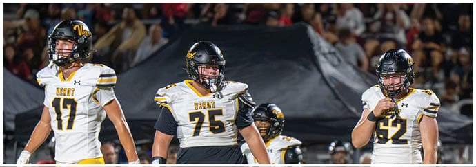 Three people wearing football uniforms and helmets stand on a field with a crowd in the background. Descripci n generada con IA