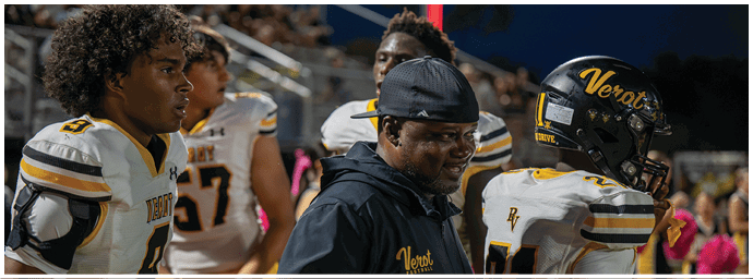 People in football uniforms and a coach wearing a cap are standing on the sidelines of a nighttime game. Descripci n generada con IA