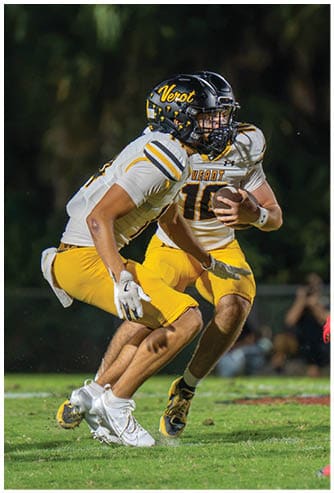 Two people in football uniforms with helmets are actively engaged in a play on a grassy field during a game. Descripci n generada con IA