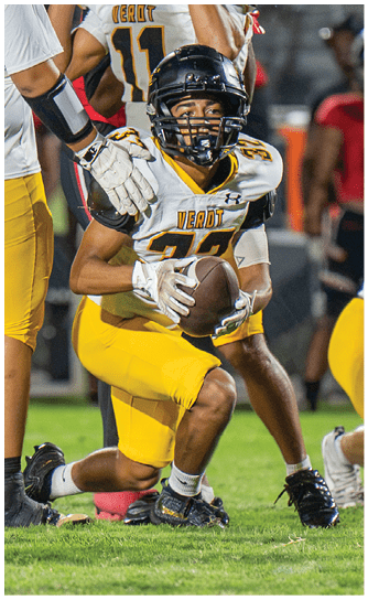 A person in a yellow and white football uniform is holding a football and preparing to make a play on a football field with other players in the background. Descripci n generada con IA