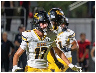 A person wearing a white and yellow football uniform with the number 17 and a black helmet is actively engaged in a game on a field with other players and blurred spectators in the background. Descripci n generada con IA