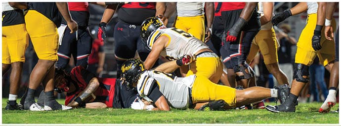Two people in yellow and white football uniforms are on the ground during a game while other players in black and yellow uniforms stand nearby on the field. Descripci n generada con IA