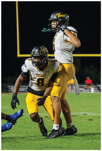 Two people in yellow and white football uniforms are on a green field at night with one person on the ground wearing a black and red uniform. Descripci n generada con IA