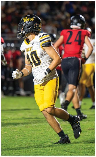 A person wearing a white and yellow football uniform with the number 40 is running on a grassy field during a game while other players in red and black uniforms are visible in the background. Descripci n generada con IA