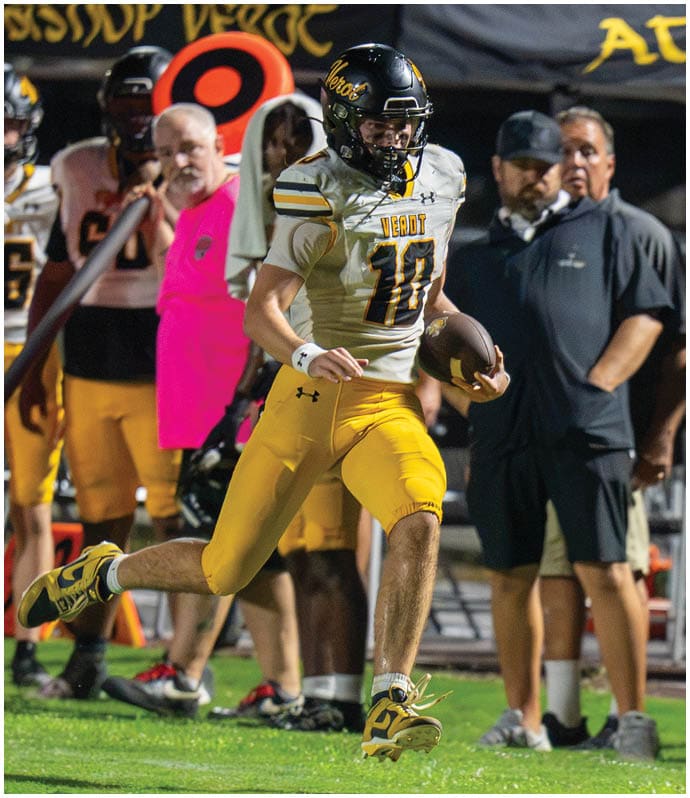 An adult wearing a football uniform with the number 10 is running on a grassy field while holding a football, with other players and coaches standing in the background. Descripci n generada con IA