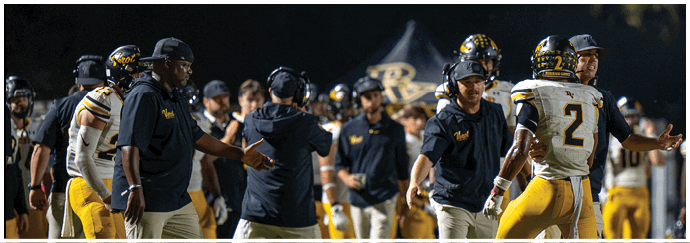 People wearing football uniforms and coaching attire are interacting on the sidelines of a football field during a game. Descripci n generada con IA